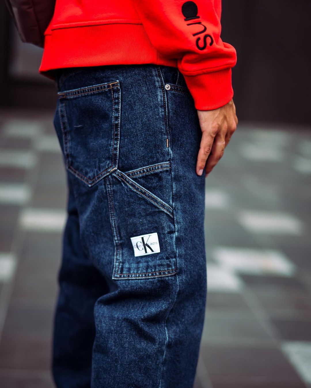 Person wearing dark blue denim work pants with a Calvin Klein logo patch on the utility pocket, paired with a red sweatshirt.