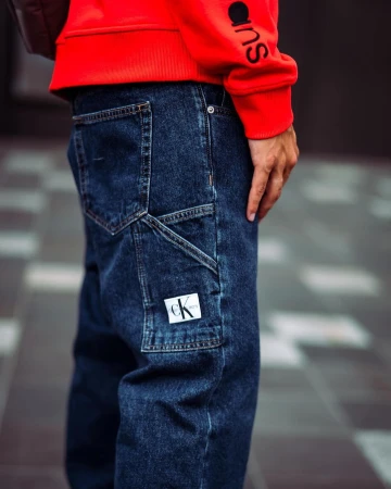 Person wearing dark blue denim work pants with a Calvin Klein logo patch on the utility pocket, paired with a red sweatshirt.