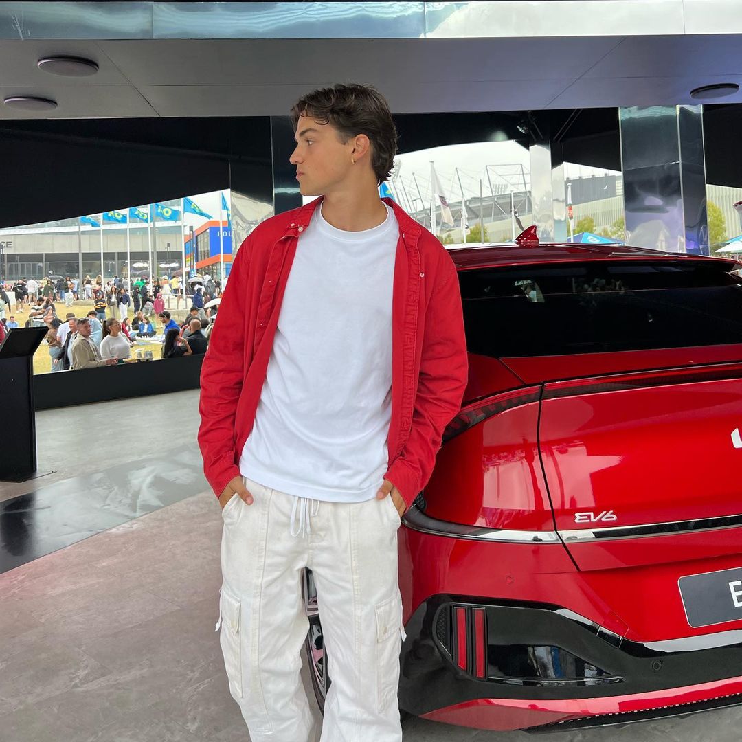 Young adult poses by a red Kia EV6 electric vehicle outside, wearing a white t-shirt, red overshirt, and white cargo pants.
