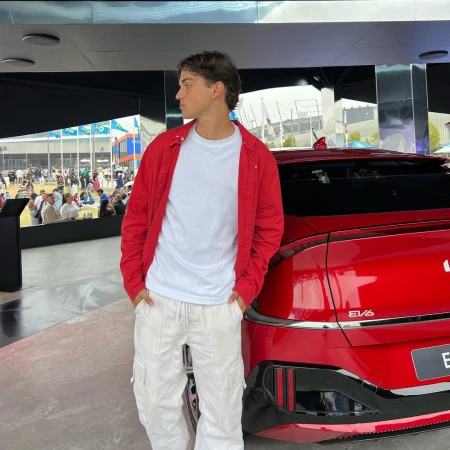 Young adult poses by a red Kia EV6 electric vehicle outside, wearing a white t-shirt, red overshirt, and white cargo pants.