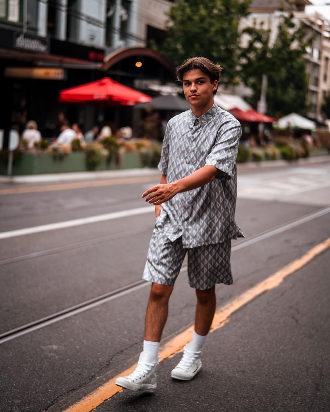 Young adult in patterned shirt and shorts walks on street, wearing high-top sneakers, with commercial buildings and red umbrellas in the background.