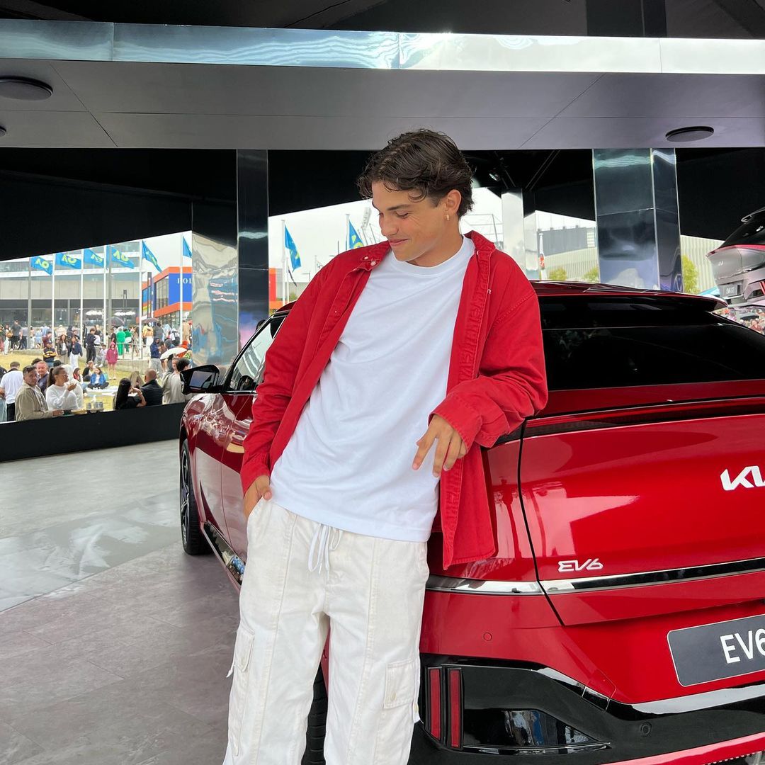 A stylish young man in a red jacket casually poses next to a vibrant red Kia EV6 car at an outdoor car show event.