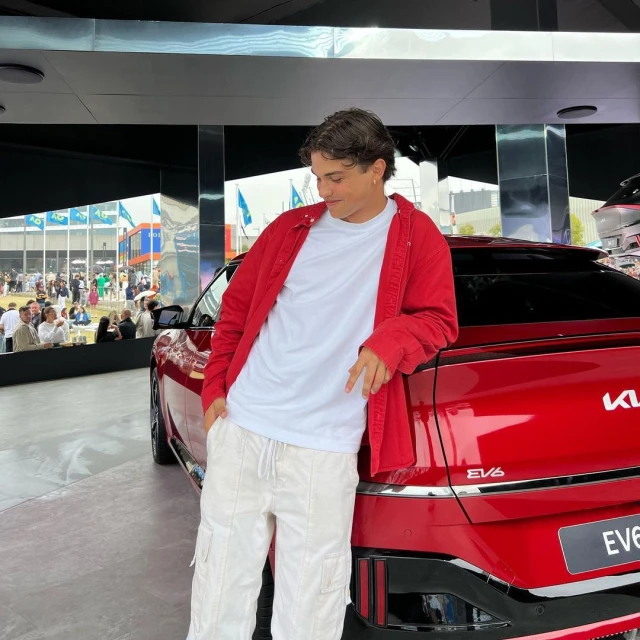 A stylish young man in a red jacket casually poses next to a vibrant red Kia EV6 car at an outdoor car show event.