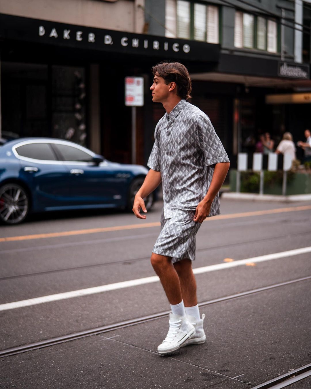 A young adult male wearing a patterned shirt and shorts set walks across a city street with a blue car and building in the background.