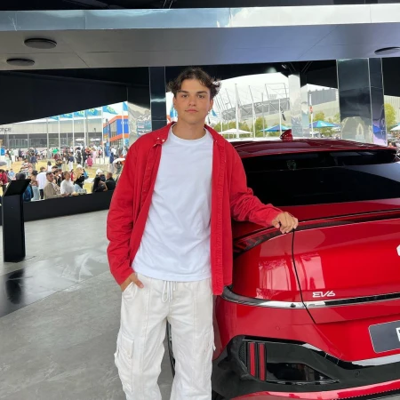 Young man in a red jacket and white cargo pants standing next to a red car in Sydney.