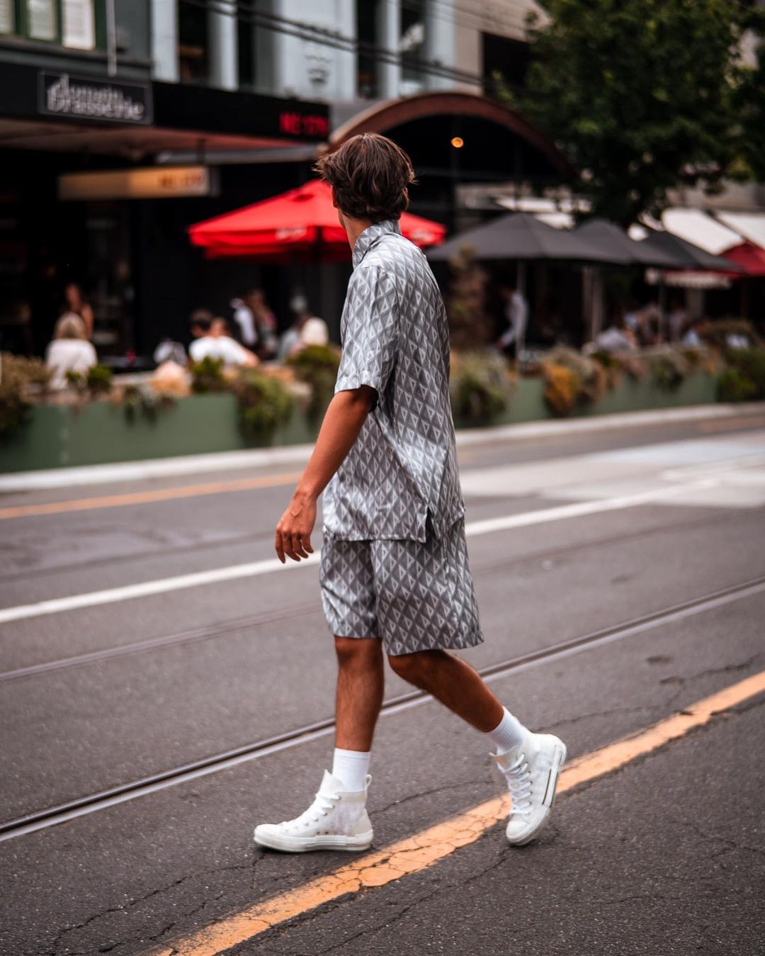 Back view of a stylish adult male in a patterned shirt and shorts walking across a crosswalk in an urban setting.