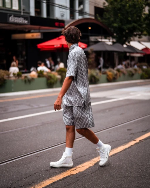 Back view of a stylish adult male in a patterned shirt and shorts walking across a crosswalk in an urban setting.