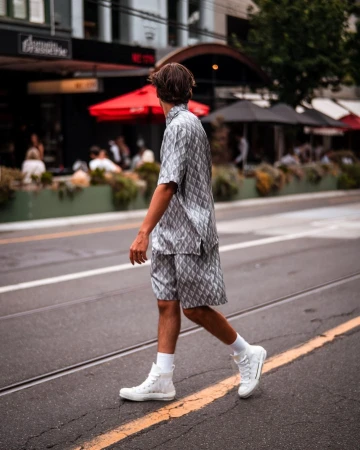 Back view of a stylish adult male in a patterned shirt and shorts walking across a crosswalk in an urban setting.