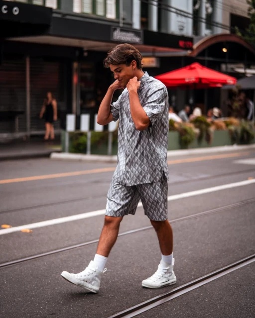 A stylish young man in a matching patterned shirt and shorts walks confidently on a city street with red awnings and buildings.