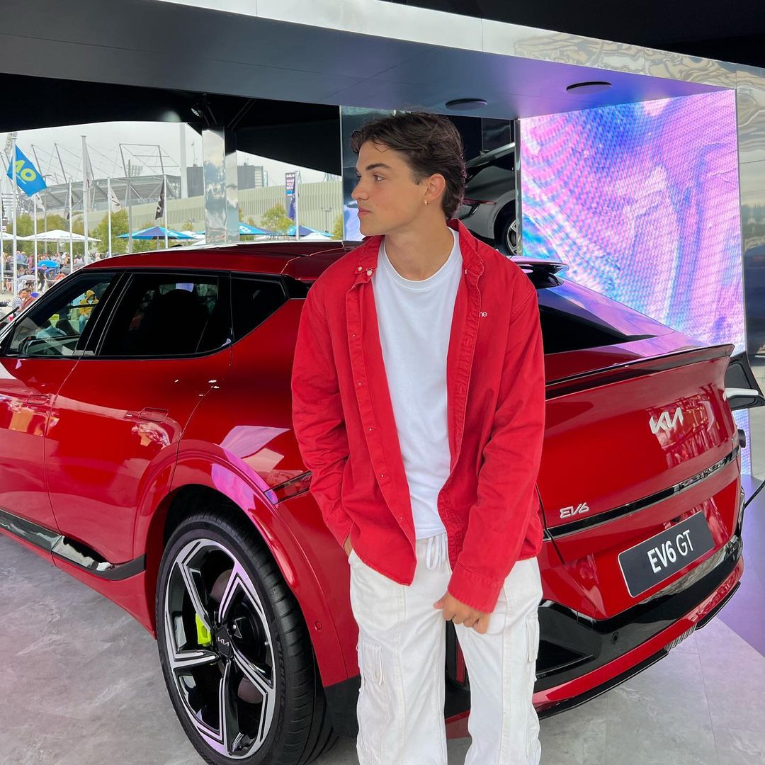 A young adult male stands in front of a red Kia EV6 GT car on display outdoors, wearing a red shirt, white t-shirt, and white pants.