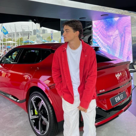 A young adult male stands in front of a red Kia EV6 GT car on display outdoors, wearing a red shirt, white t-shirt, and white pants.