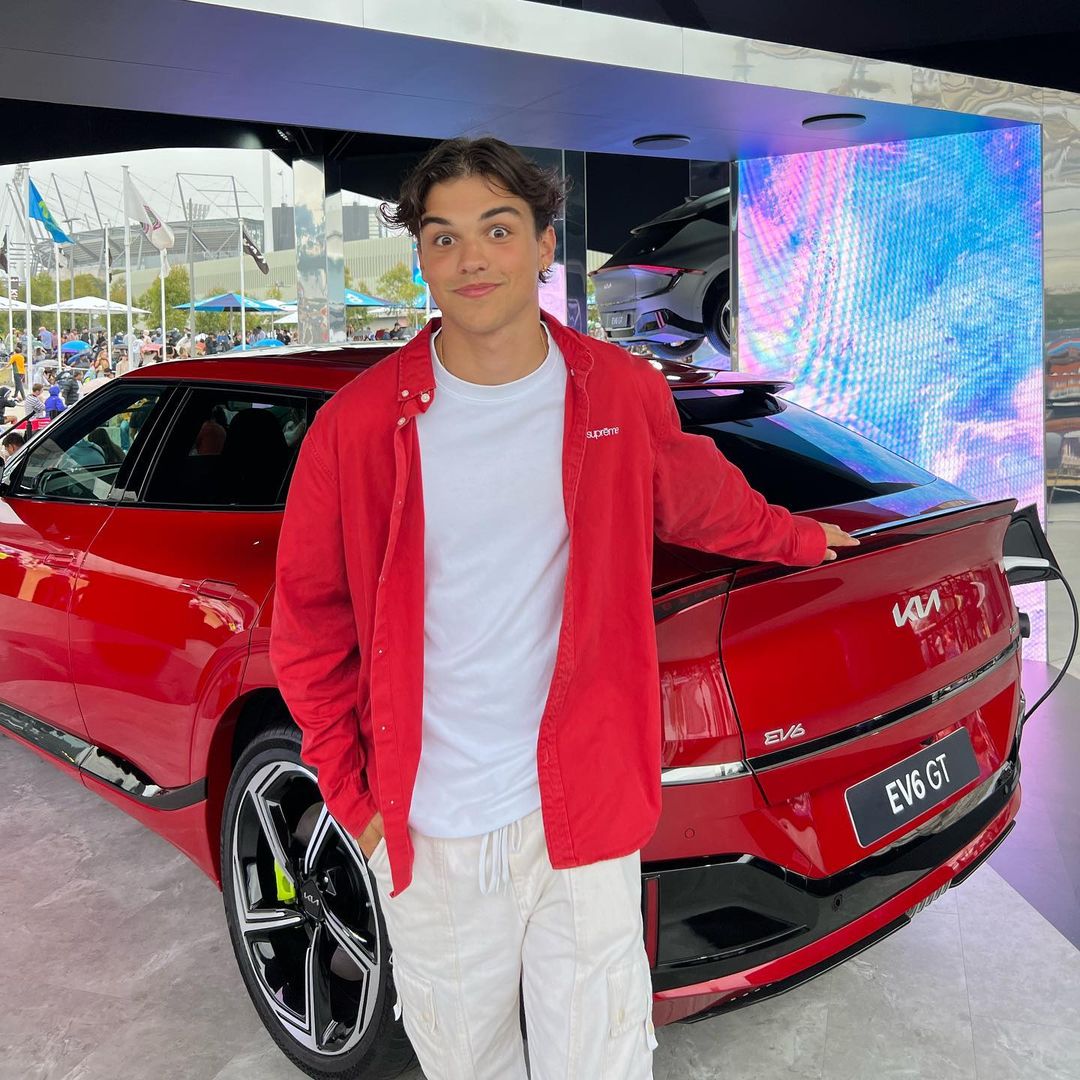 A young adult male poses next to a red Kia EV6 GT electric car at an automotive event, with a bright LED screen in the background.