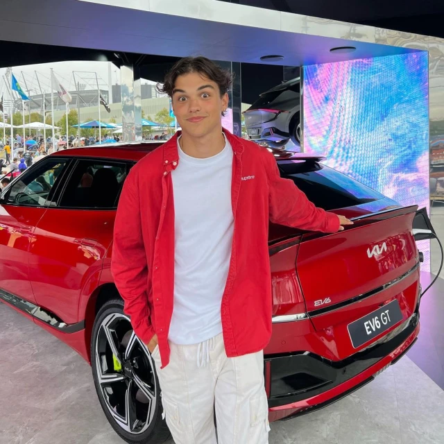 A young adult male poses next to a red Kia EV6 GT electric car at an automotive event, with a bright LED screen in the background.