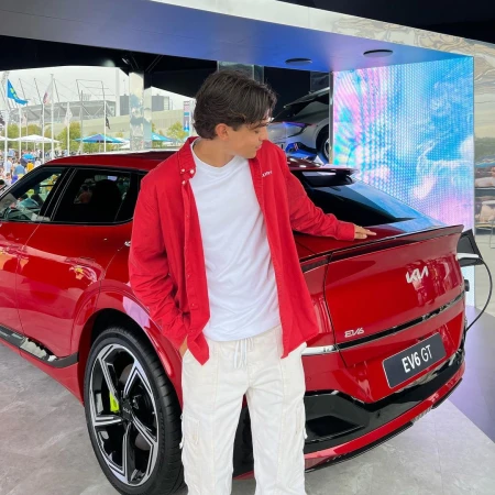 Young man in a red shirt and white pants stands next to a red Kia EV6 GT electric vehicle, showcasing modern automotive design and electric technology.