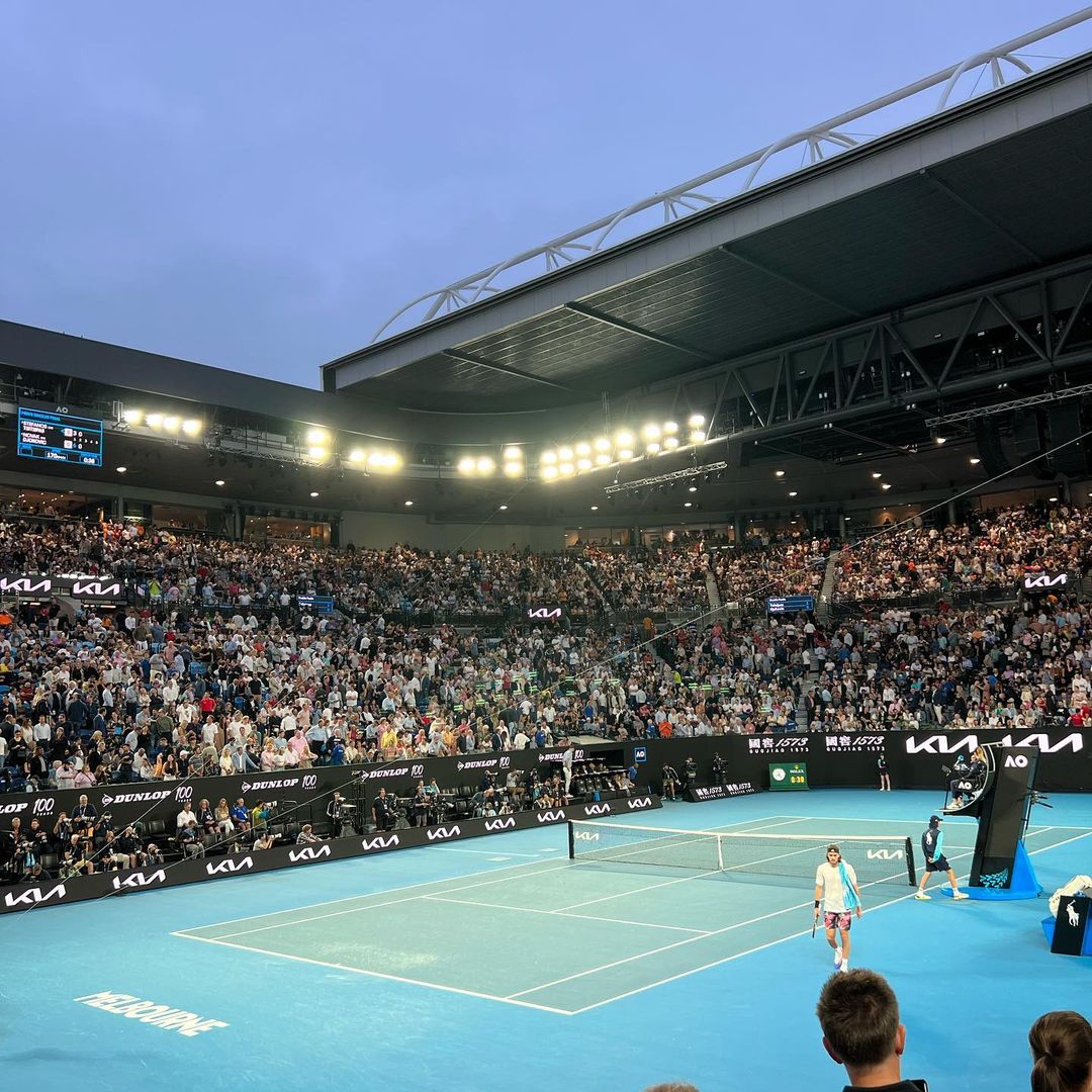 A wide shot of a tennis match being played in a packed stadium with spectators in the stands and bright lighting overhead.