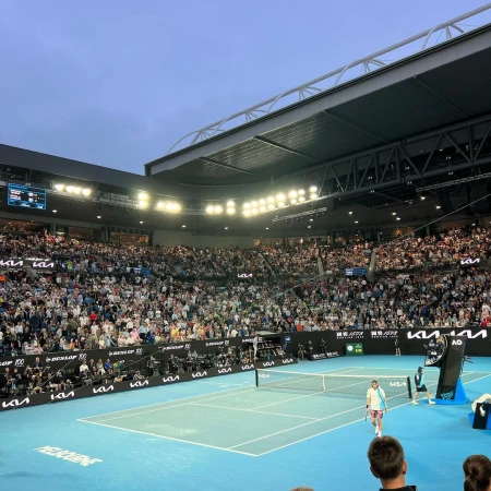 A wide shot of a tennis match being played in a packed stadium with spectators in the stands and bright lighting overhead.