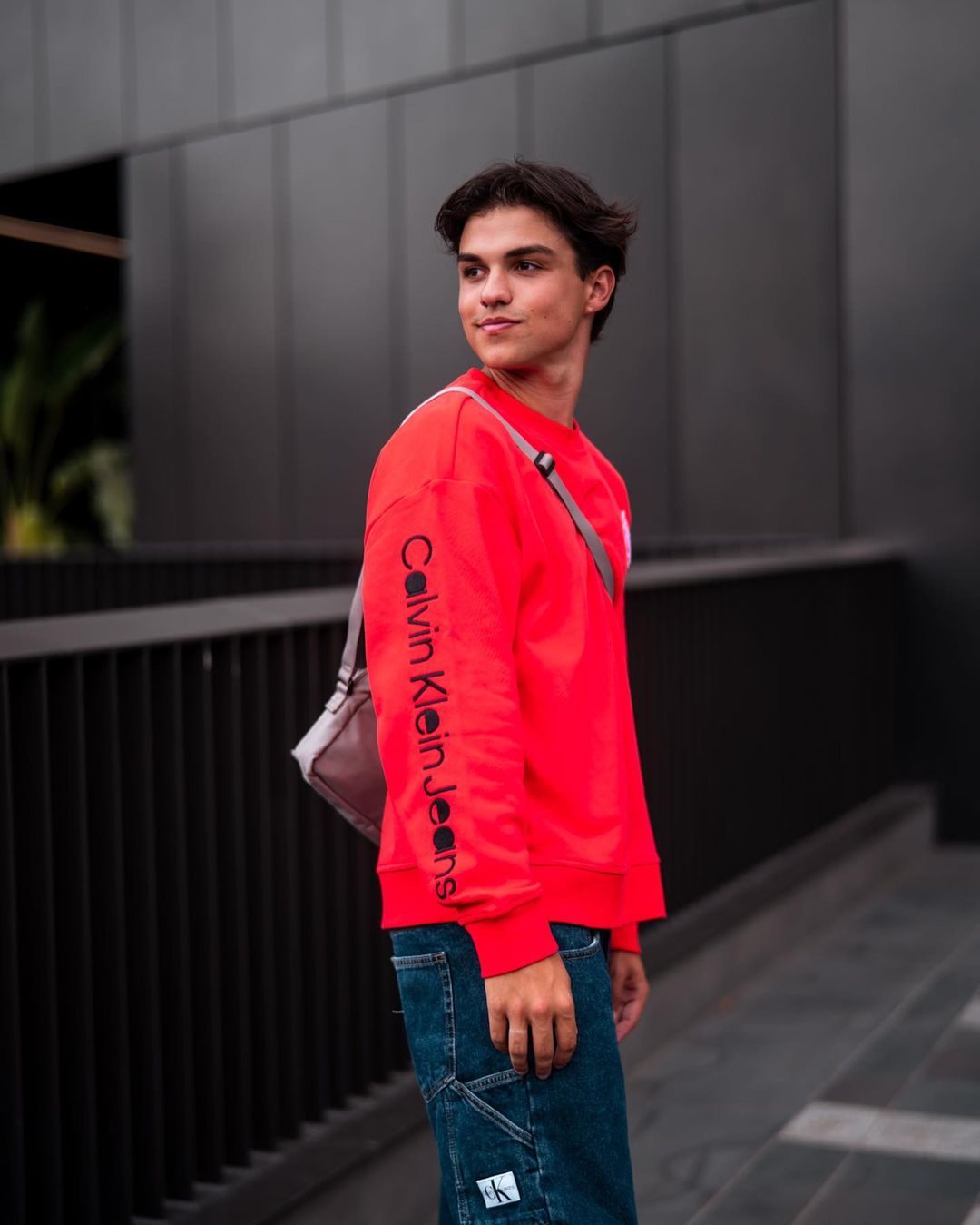 Teenage male model in a red Calvin Klein sweatshirt and jeans poses near a modern grey building with natural light and soft shadows.