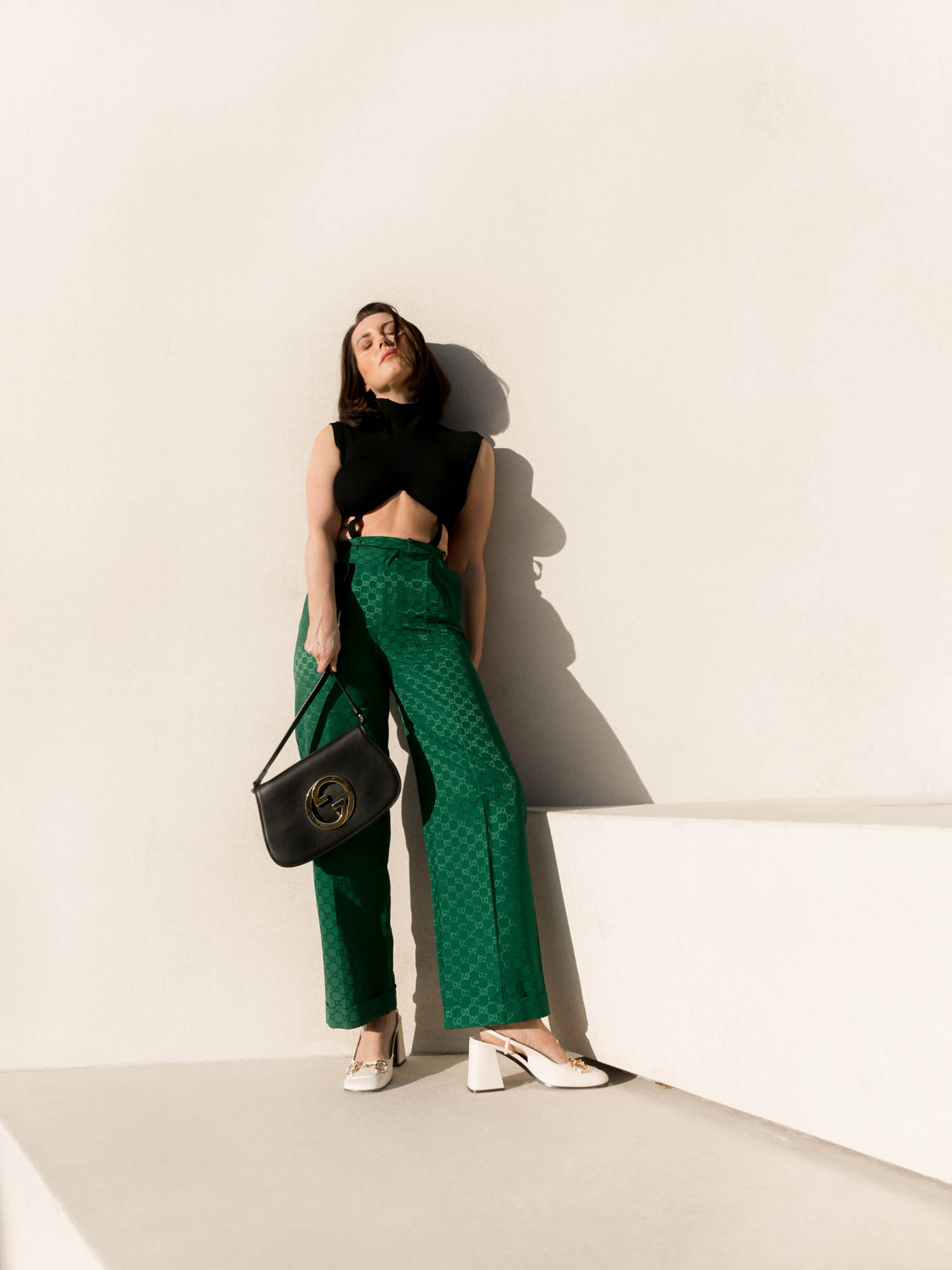 Fashion portrait of a woman in a black top and green pants leaning against a white wall, holding a black purse, and wearing white shoes.