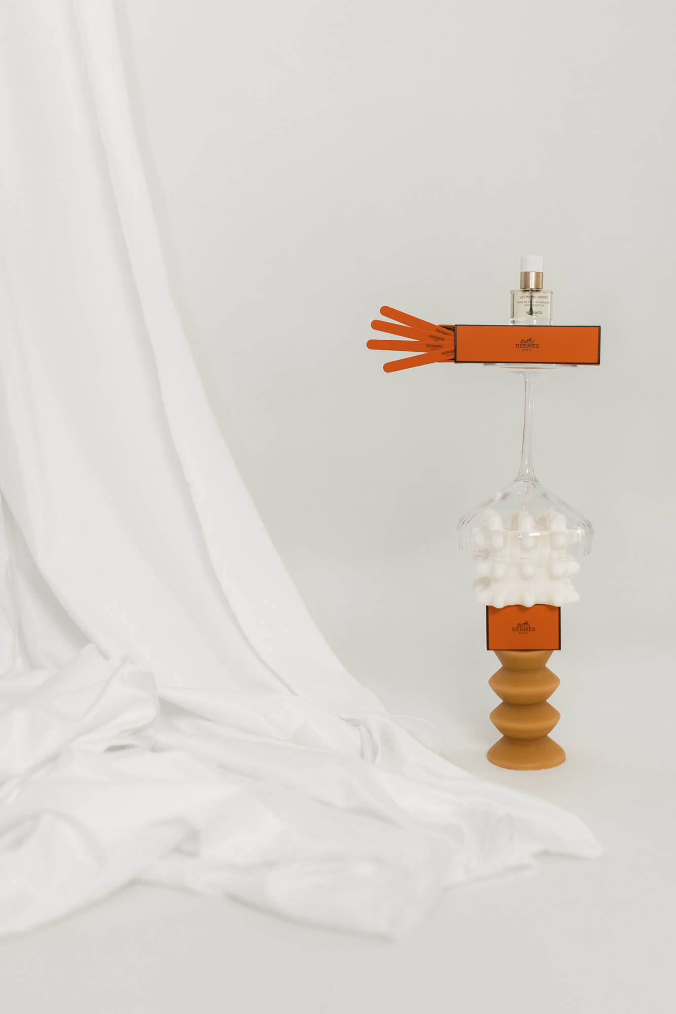 Still life featuring a bottle of perfume on top of a glass and wooden stand, beside a white fabric, in a minimalist composition.