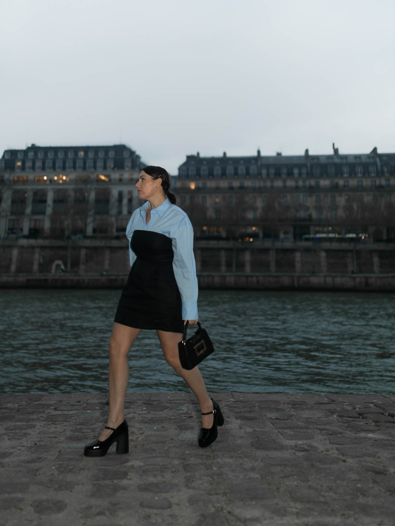 Woman in a blue shirt and black dress walks along a stone path next to a river in a city, carrying a black purse and wearing platform shoes.