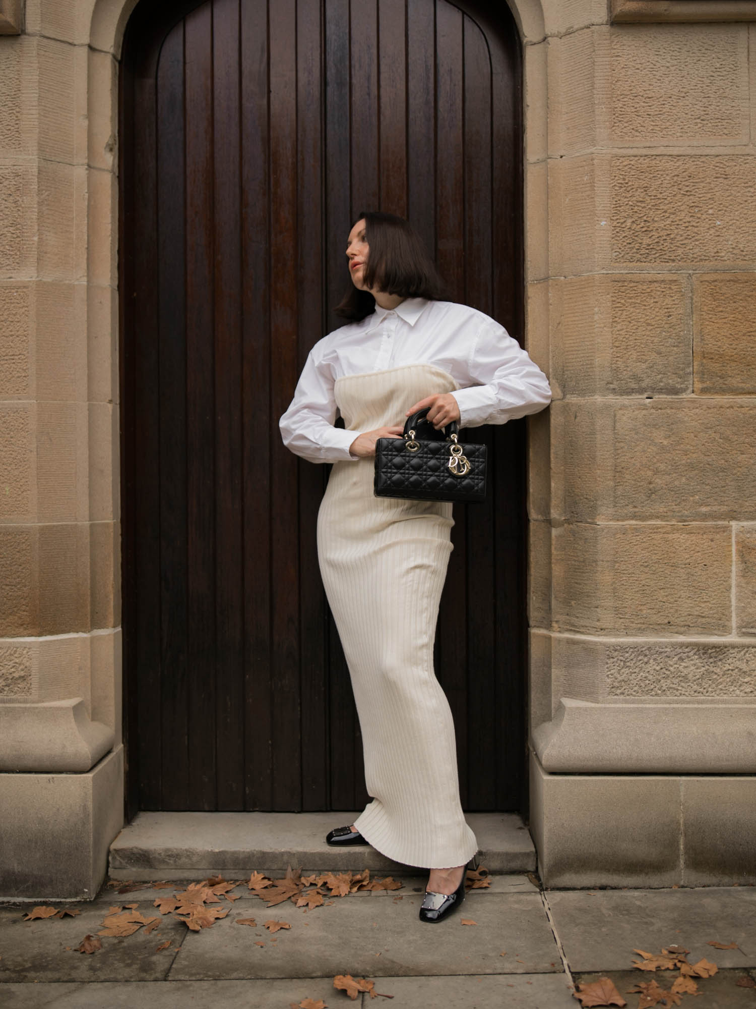 Woman in a white shirt and beige dress stands in front of a dark brown door and light stone wall while holding a black purse.