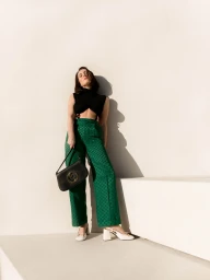 Woman in black top and green patterned trousers leaning against a white wall holding a black handbag.