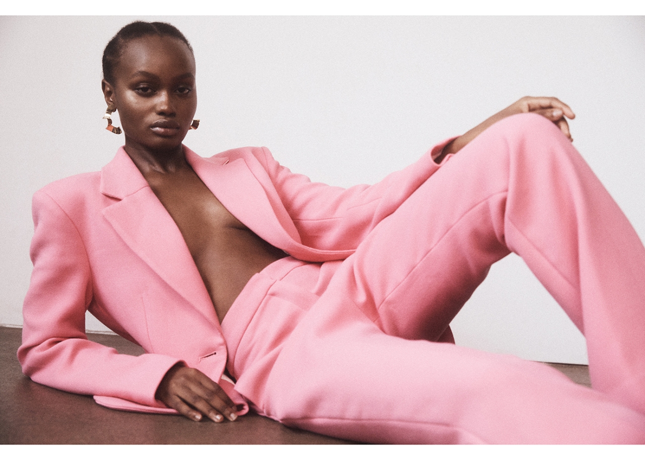 Elegant Black woman in a pink blazer and wide-leg pants laying down indoors for a fashion shot, highlighted by studio lighting.