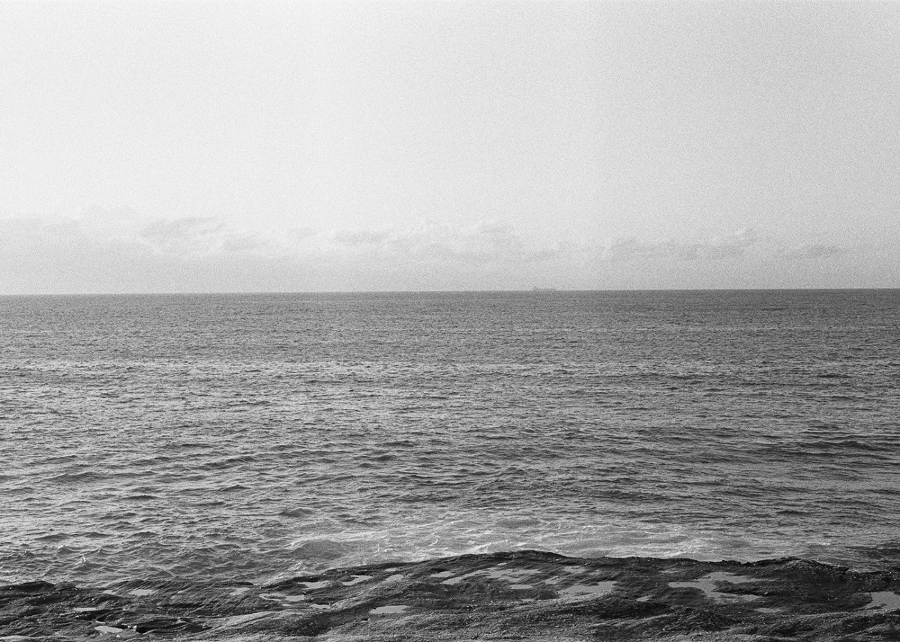 Monochromatic seascape featuring calm ocean waters under a bright sky, with rocky textures in the foreground and a distant ship visible.
