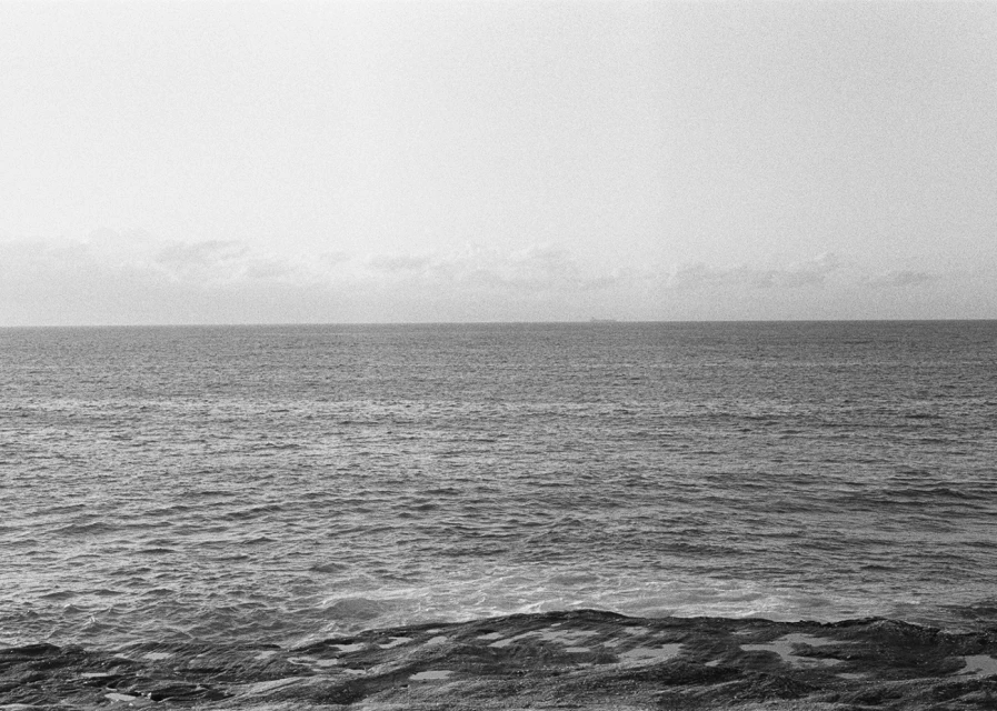 Monochromatic seascape featuring calm ocean waters under a bright sky, with rocky textures in the foreground and a distant ship visible.