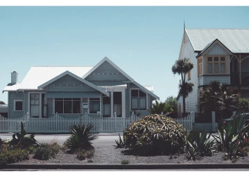 Exterior shot of a single-story light blue house with a white picket fence, corrugated metal roof, and lush garden plants in the foreground.