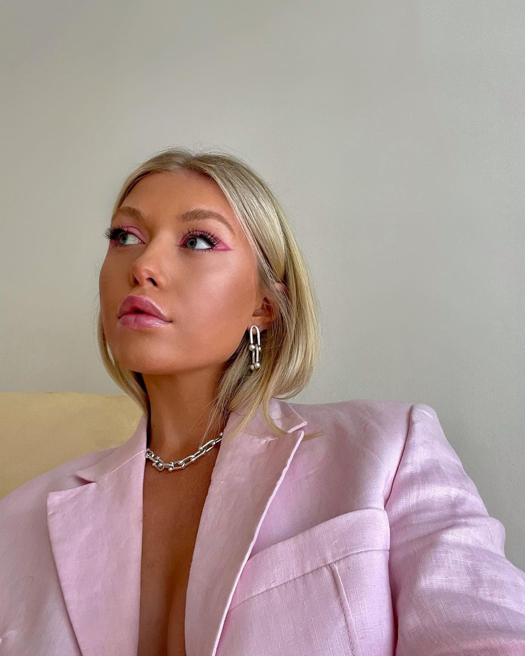 Blonde woman in a pink blazer and statement jewelry poses in a close-up studio shot with pink eyeliner.