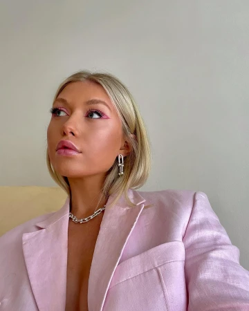 Blonde woman in a pink blazer and statement jewelry poses in a close-up studio shot with pink eyeliner.