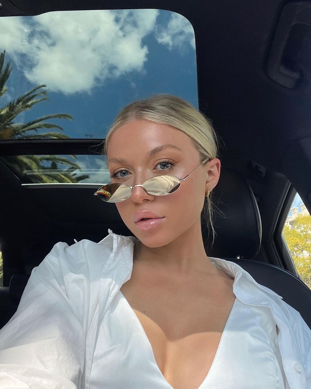Close-up of a blonde woman in a car with an open sunroof, wearing sunglasses and a white shirt, with blue sky and palm trees visible.