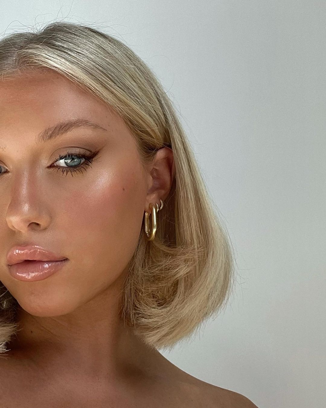 Close-up beauty shot of a blonde adult woman with shoulder-length hair, winged eyeliner, and nude lip gloss, wearing gold hoop earrings.