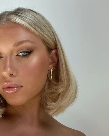 Close-up beauty shot of a blonde adult woman with shoulder-length hair, winged eyeliner, and nude lip gloss, wearing gold hoop earrings.