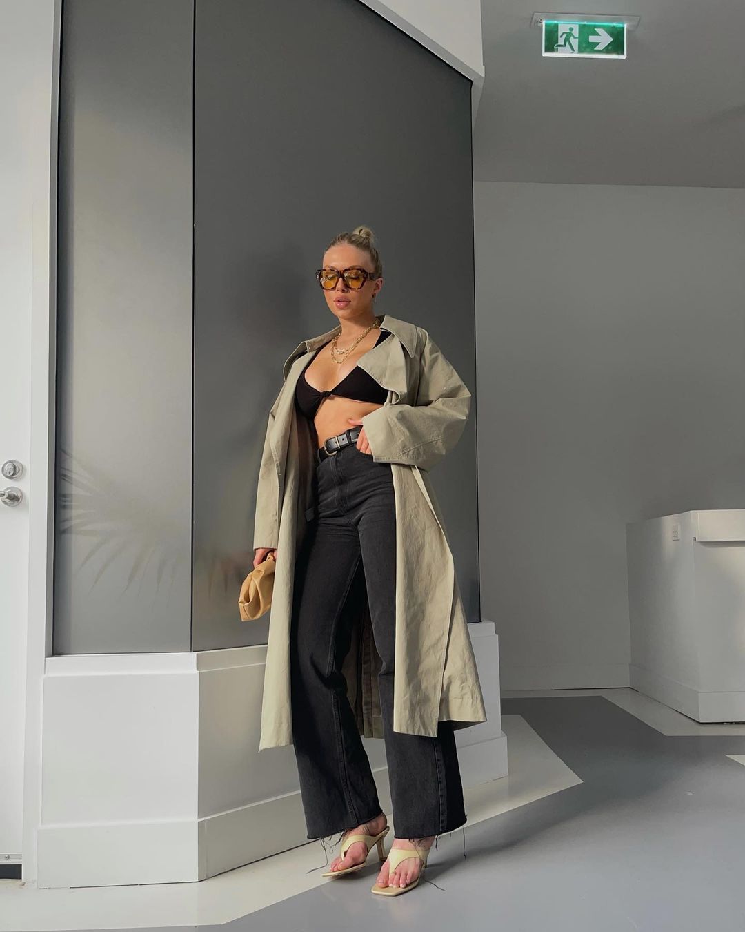 Woman in trench coat, black bra top, and jeans poses in an indoor urban setting with neutral colors, holding a small beige clutch.