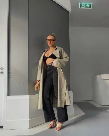 Woman in trench coat, black bra top, and jeans poses in an indoor urban setting with neutral colors, holding a small beige clutch.