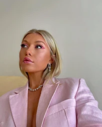 Close-up portrait of a blonde woman wearing a pink blazer, silver chain necklace, and drop earrings against a plain wall.