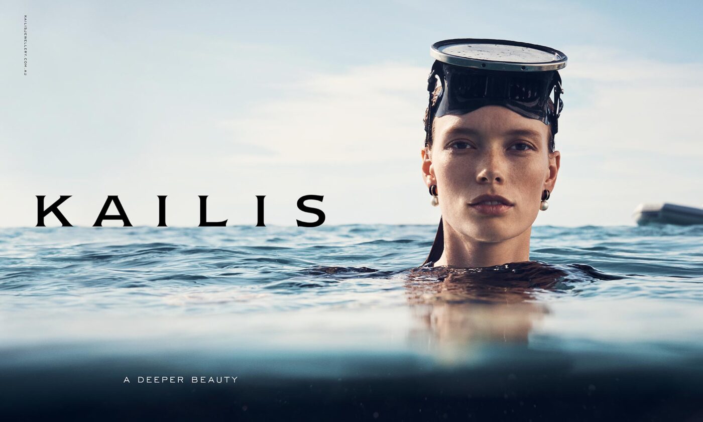 A woman with a black diving mask is partially submerged in blue water, against a bright sky, in a promotional beauty shot.