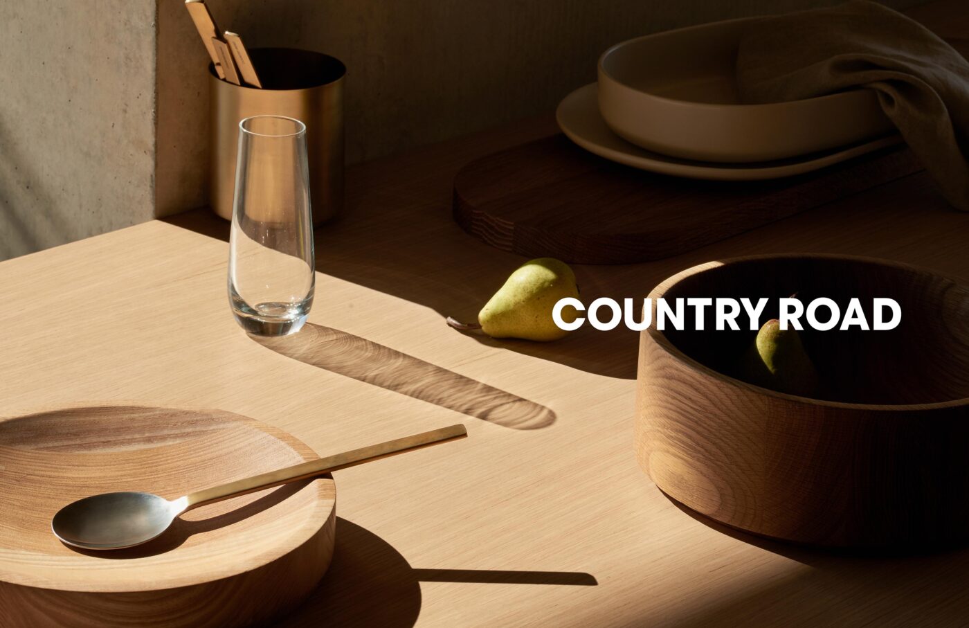 Still life of wooden bowls, a glass vase, pear, and metal cutlery displayed on a table with neutral tones and shadows. Tabletop product photography.
