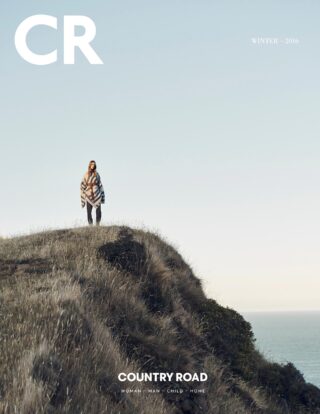 Fashion editorial featuring a woman standing on a grassy hill overlooking the ocean, wearing a poncho and pants against a sky background.