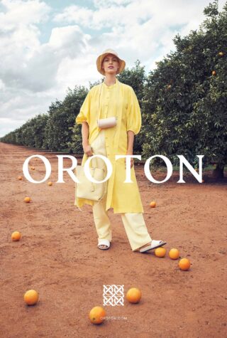 A woman in a yellow outfit and straw hat stands in an orange grove for a fashion advertisement, with oranges scattered on the ground.