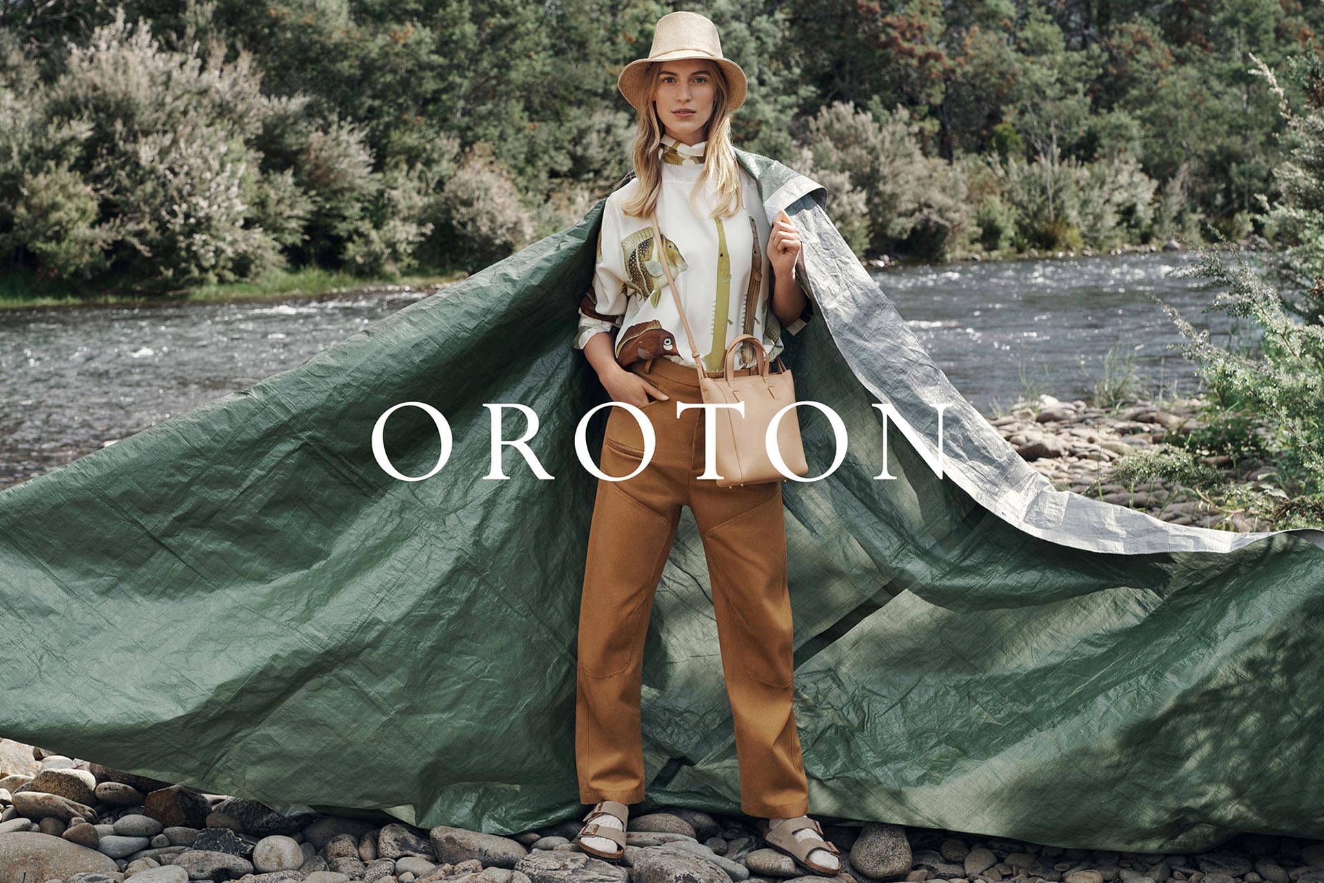 A blonde woman in a hat and patterned shirt with a purse poses in front of a river with a large tarp draped behind her.