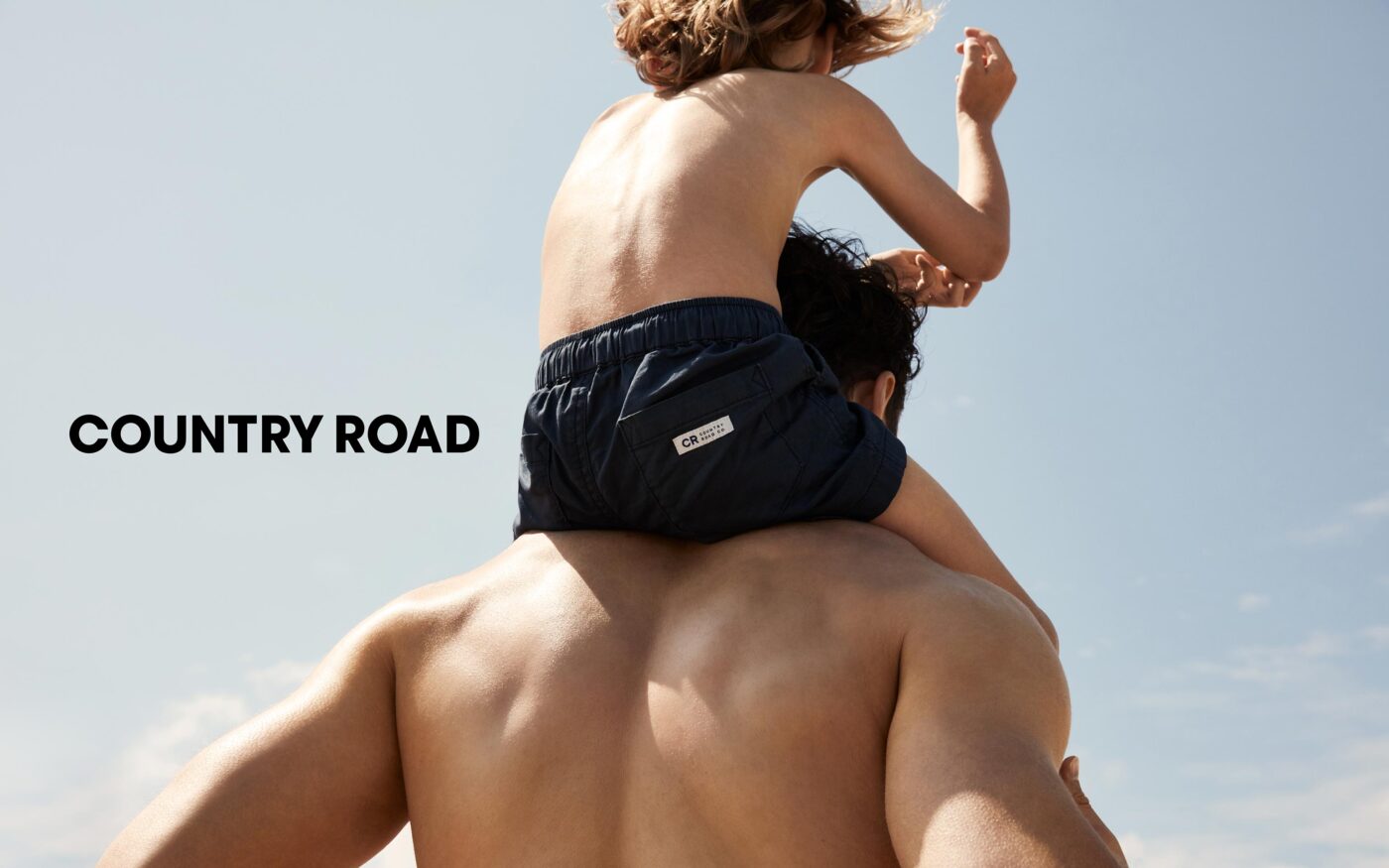 Father and child in beachwear on a sunny day with a clear sky. The child sits on the father's shoulders, and 'Country Road' text is visible.
