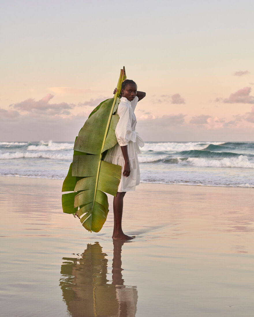 A woman poses barefoot on a sandy beach at dusk wearing a white dress and carrying a large, broad green banana leaf.