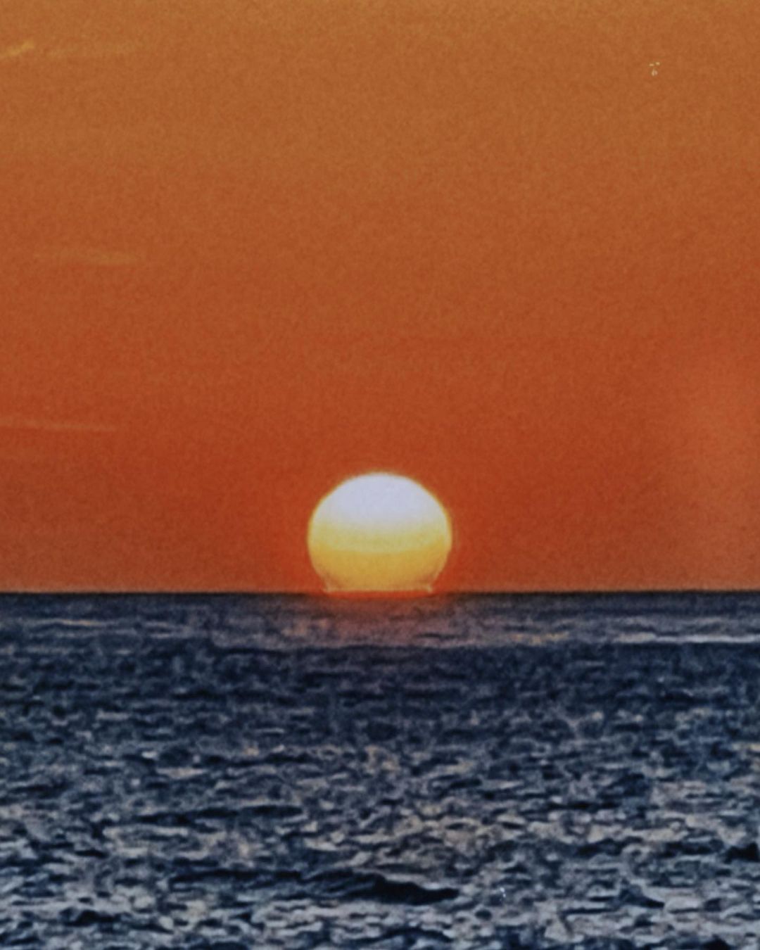 Sunset over the ocean with the sun appearing as a bright sphere touching the water, creating a horizon line. Sky is orange-red gradient.