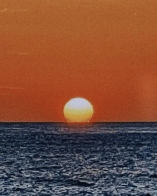 Sunset over the ocean with the sun appearing as a bright sphere touching the water, creating a horizon line. Sky is orange-red gradient.