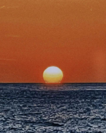 Sunset over the ocean with the sun appearing as a bright sphere touching the water, creating a horizon line. Sky is orange-red gradient.
