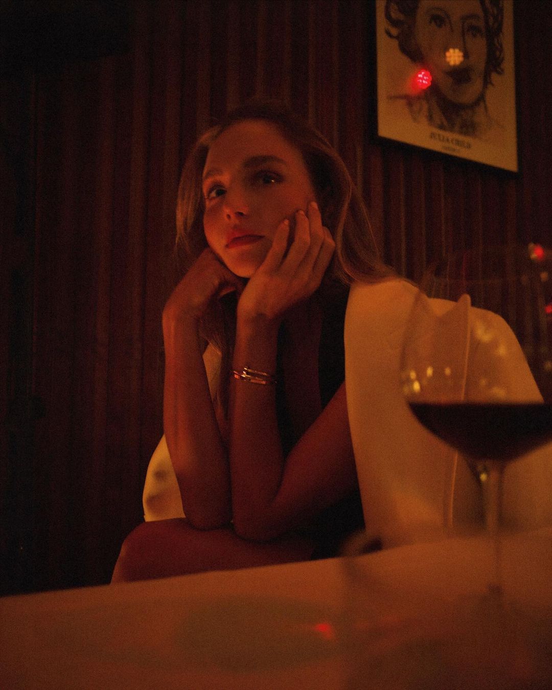 Woman sits at a table in warm light, looking pensive with a wine glass in view and a framed sketch hanging on the paneled wall.
