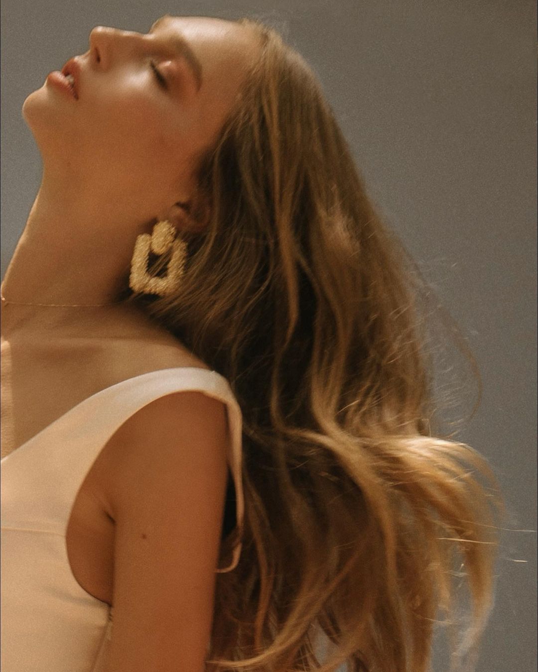 Blonde woman in a sleeveless white top, poses indoors with eyes closed. She wears statement square earrings, with grey backdrop.